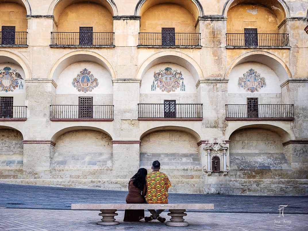 Pablo and Seraphine sitting together on a bench, small against the tall arcaded façade behind them.