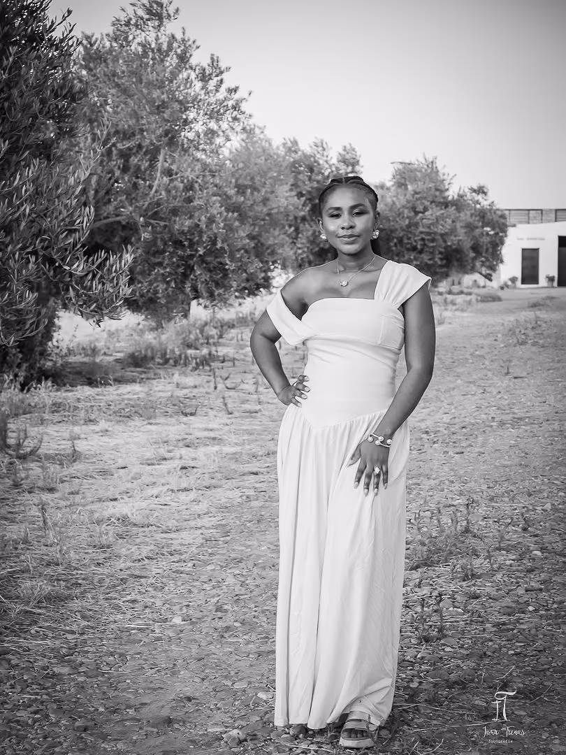 Seraphine standing in a long draped white gown on a path through an olive grove, in black and white.