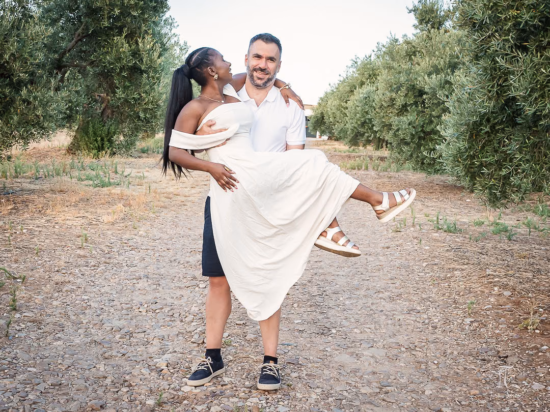 Pablo lifting Seraphine off her feet on a gravel path between olive trees, both laughing.