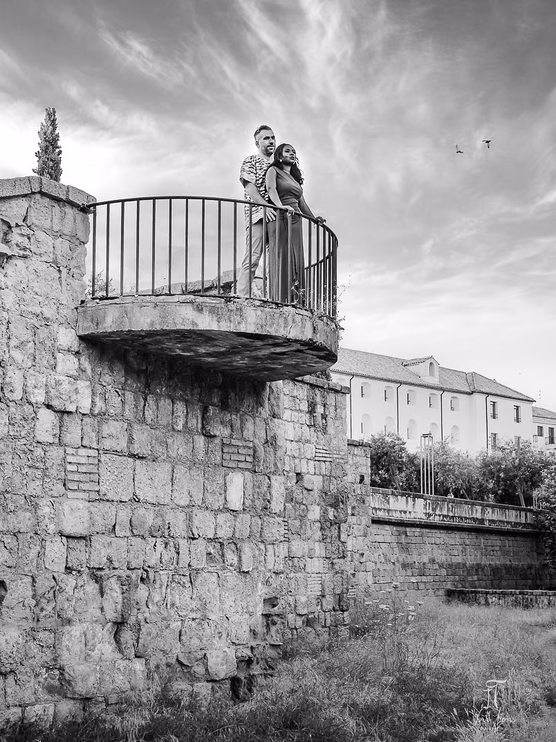 Pablo and Seraphine standing on a stone balcony of the Roman Bridge in Córdoba, in black and white against a dramatic sky.