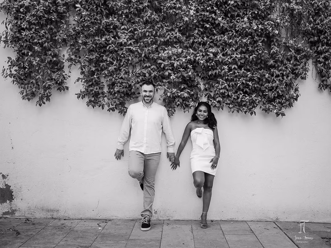 Pablo and Seraphine walking hand in hand along a whitewashed wall under a dark hedge, in black and white.